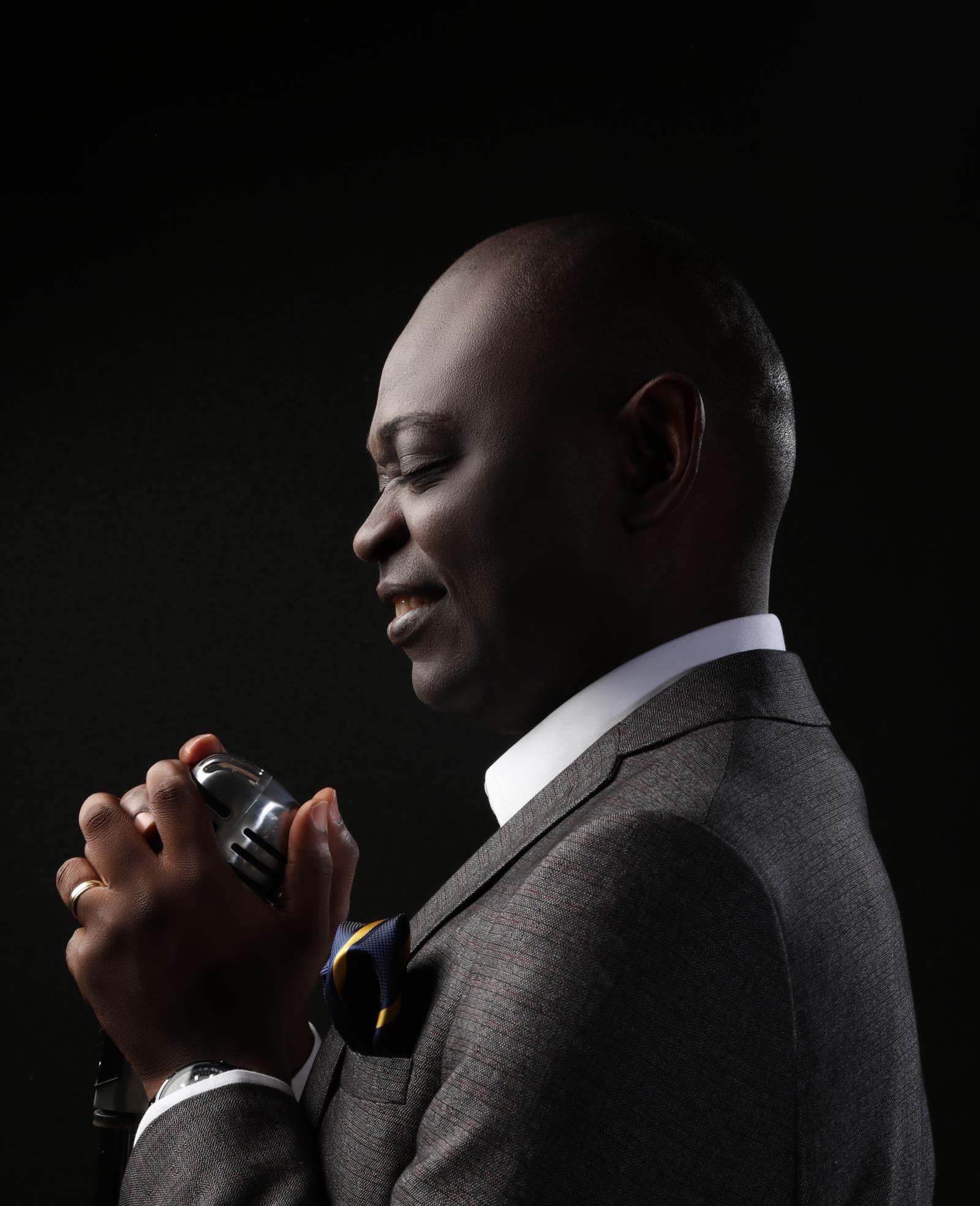 Yemi Alafifuni singing into a vintage microphone, lit from one side against near-black