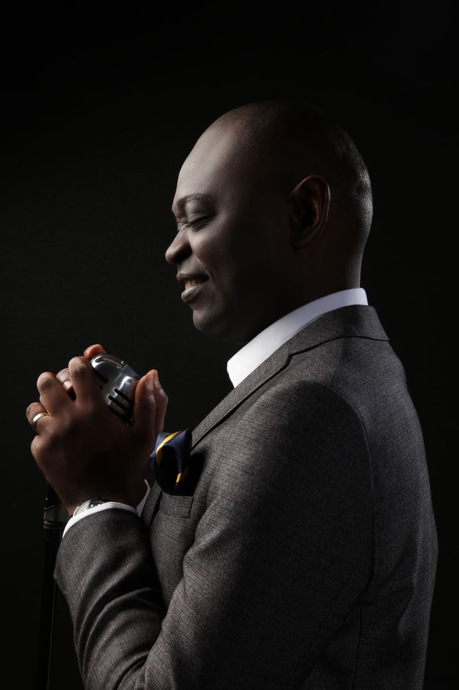 Yemi Alafifuni in profile, singing into a vintage microphone, lit from one side against black