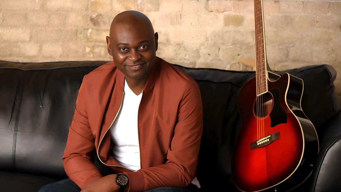 Yemi Alafifuni seated on a leather sofa beside an acoustic guitar, brick wall behind